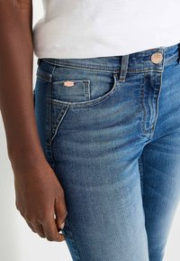 Jeans de mezclilla en un lavado intermedio con ligero desgastes, que cuentan con herrajes de cobre, un bolsillo frontal y una pequeña etiqueta de marca metálica en la cinturilla.