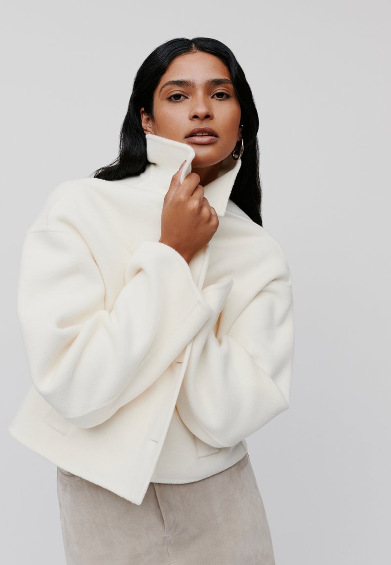 Witte fleecejack met een hoge kraag en oversized mouwen, gedragen over een beige kledingstuk, met een minimalistisch ontwerp en een gladde textuur.