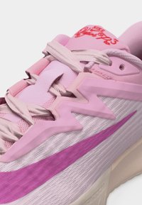 Gros plan sur une chaussure Nike rose et violette montrant une superposition stylisée, des lacets, un tissu en maille et un logo partiel sur le côté et la languette.