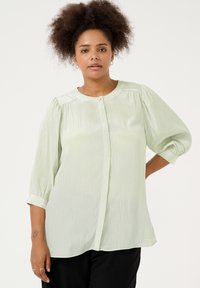 Blusa verde chiaro, semi-trasparente, con finitura testurizzata, scollatura tonda, chiusura a bottoni e maniche a sbuffo. Tessuto morbido e fluido.