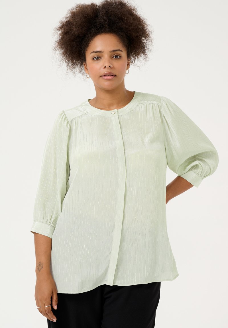 Blusa verde chiaro, semi-trasparente, con finitura testurizzata, scollatura tonda, chiusura a bottoni e maniche a sbuffo. Tessuto morbido e fluido.