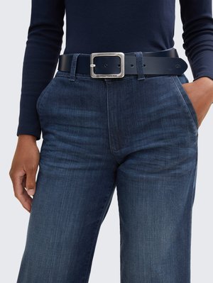 Marinefarbener Langarmtop kombiniert mit dunkelblauen Jeanshosen. Schwarzer Gürtel mit rechteckiger silberner Schnalle, 2 sichtbare Taschen und einem strukturierten Stoff.