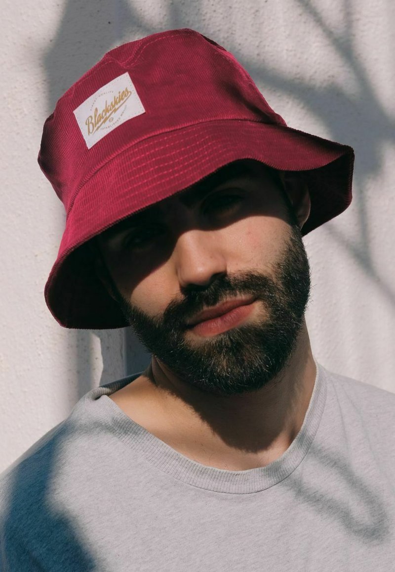 Maroonin värinen ämpärihattu, jossa on tekstuurimateriaali ja valkoinen logo-patch. Yhdistetty harmaaseen paitaaan, neutraalin taustan kanssa.