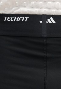 Ceinture noire de short de sport avec texte blanc « TECHFIT » et logo Adidas à trois bandes sur tissu blanc texturé en haut.