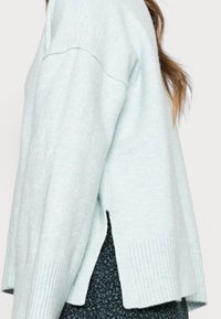 Pull tricoté bleu clair avec poignets côtelés, fentes latérales et coupe décontractée. Texture douce, finition légèrement chinée et coutures épaules tombantes.