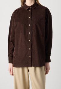 Camicia di velluto a coste marrone con colletto, maniche lunghe e chiusura anteriore con bottoni. Presenta una tasca sul petto e bottoni beige.