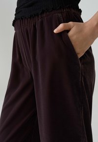Brown, velvet lounge pants with an elastic waistband, side pockets, and a relaxed fit, featuring a smooth texture and subtle sheen.