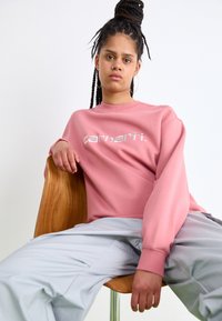 Rosa sweatshirt med hvidt brandlogo, løs pasform, ribbede manchetter, parret med lys grå bukser, siddende på en træstol.