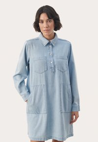 Svetlo modra denim obleka s kragnatim izrezom, gumbom na sprednji strani, dvema žepoma na prsih in dvema velikima žepoma na straneh. Dolgi rokavi in sproščen kroj.