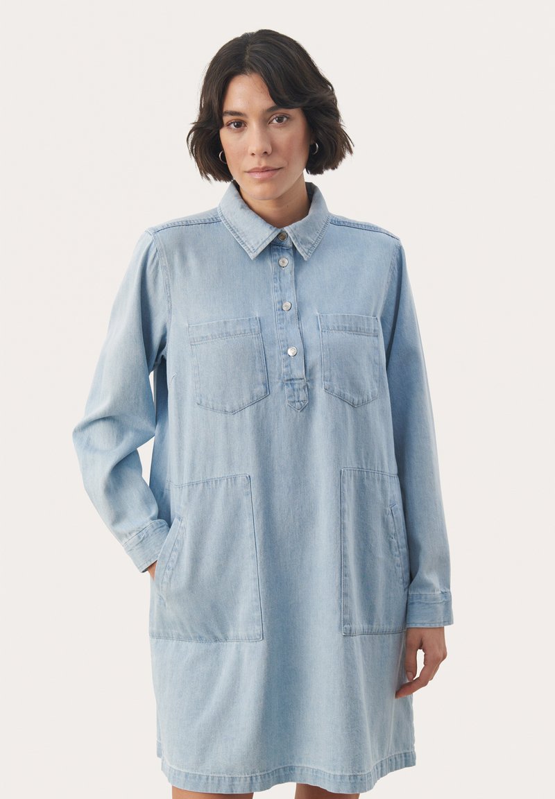 Svetlo modra denim obleka s kragnatim izrezom, gumbom na sprednji strani, dvema žepoma na prsih in dvema velikima žepoma na straneh. Dolgi rokavi in sproščen kroj.