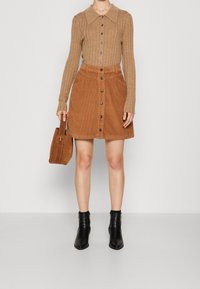 Suéter marrón de canalé con cuello, combinado con una falda de pana beige con botones en la parte delantera. Botines negros y un bolso marrón completan el look.