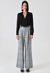 Zwarte blouse met ruches, gecombineerd met hoge taille, brede grijze geplaid broek. Zwarte schoenen en zilveren sieraden accentueren de outfit.