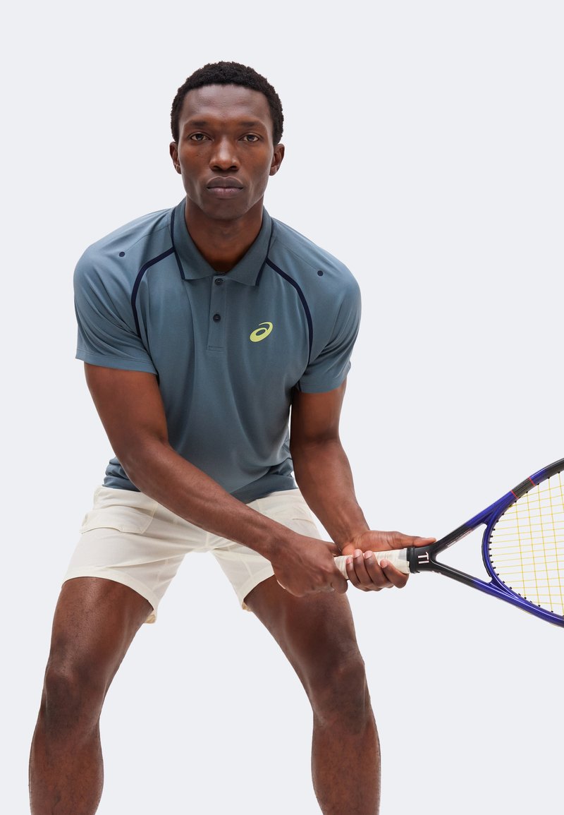 Männlicher Tennisspieler in blauem Polo und weißen Shorts hält den Schläger mit beiden Händen und bereitet einen Schlag vor vor einfachem Hintergrund.