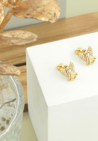 Orecchini a forma di farfalla in oro con accenti di gemme trasparenti e un dettaglio a forma di cuore in argento, esposti su un piedistallo bianco con un vaso di vetro nei dintorni.