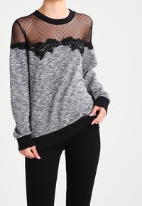 Sudadera de punto gris con una sisa de malla negra semitransparente con borde de encaje, puños y dobladillo negros, llevada sobre pantalones negros.