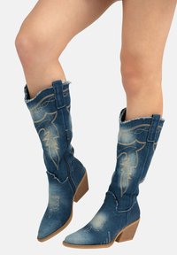 Botas de vaquero de denim con punteras puntiagudas, costuras doradas decorativas y un borde superior ligeramente deshilachado. Equipadas con una cremallera lateral y un tacón apilado.
