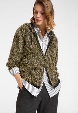 Hoodie zippé vert olive, texturé, avec des bordures et des cordons noirs, porté par-dessus une chemise rayée blanche et noire avec des manches trois-quarts.