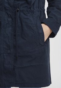 Tumši zilas krāsas viegla jaka ar saitītēm jostasvietā, sānu kabatām un teksturētu, kroku audumu virsmas apdari.