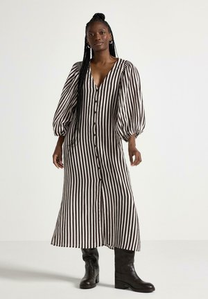 Donna in piedi che indossa un vestito a righe verticali bianche e nere con bottoni, maniche a sbuffo e stivali neri alti fino al ginocchio.