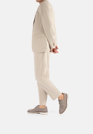 Beige maßgeschneiderter Anzug mit einreihiger Jacke und passenden Hosen. Graue Wildlederschuhe mit weißen Sohlen und braunen Schnüren. Glatte Textur.