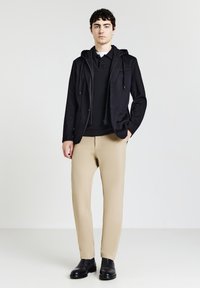 Schwarze Zip-Jacke mit Kapuze, über einem schwarzen Pullover und beigen Hosen, kombiniert mit schwarzen Schuhen, vor einem schlichten Hintergrund.