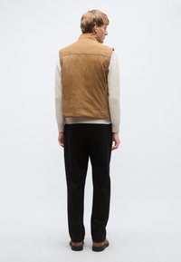 Semiš brezrokavnik svetlo rjave barve z visokim ovratnikom, nošen preko krem puloverja. Črne hlače in rjava obutev dopolnjujeta oblačilo.