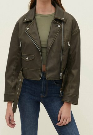 Woman wearing a dark olive cropped leather jacket with silver zippers, paired with a green crop top and blue denim jeans.
