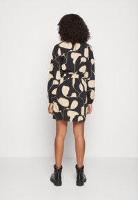 Vestido preto com padrão abstrato bege, mangas longas, cintura ajustada e fecho nas costas. Usado com botas pretas de tornozelo.