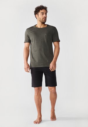 Mann steht barfuß und trägt ein dunkelgrünes kurzärmeliges Hemd mit Brusttasche sowie schwarze, knielange Shorts vor weißem Hintergrund.