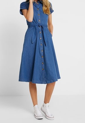 Femme portant une robe midi en denim bleu à boutons, ceinturée à la taille, et des baskets montantes blanches, debout devant un fond uni.