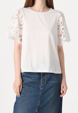 Camiseta blanca de algodón con mangas cortas de encaje que presentan patrones florales. Cuello redondo y ajuste relajado, combinada con una falda vaquera.