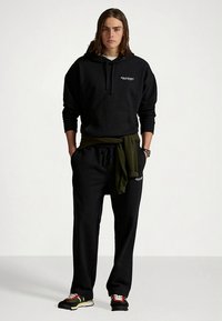 Svart hoodie och joggingbyxor i bomull, med en grön tröja knuten runt midjan. Har logotypdetaljer och sportiga sneakers.