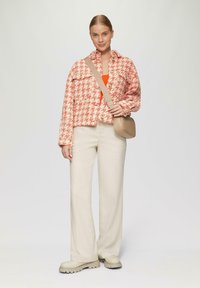 Rood en crèmekleurig houndstooth-jack, nauwsluitende beige broek, crèmekleurige schoenen met dikke zolen en een kleine beige schoudertas. Oranjetop eronder.