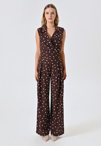 Jumpsuit marrone senza maniche con pois bianchi, caratterizzato da un design avvolgente, gambe ampie e pieghe in vita. Tessuto dalla texture liscia.