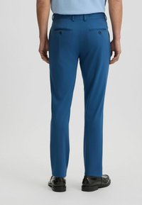 Blauwe op maat gemaakte broeken met een soepele textuur, voorzien van twee achterzakken met zwarte knopen en een slim fit ontwerp, gedragen met zwarte schoenen.
