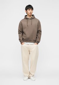 Νεαρός άνδρας στέκεται με τα χέρια στις τσέπες, φορώντας καφέ φόρμα με κουκούλα, ανοιχτόχρωμο καρό πουκάμισο, εκρού παντελόνι με φαρδύ πόδι και λευκά αθλητικά παπούτσια.