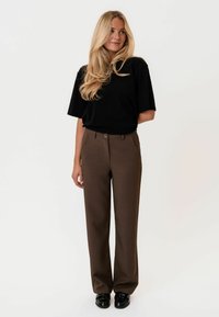 Černé oversized tričko spárované s hnědými, upravenými kalhotami s knoflíkovým zapínáním a rovnými nohavicemi. Černé mokasíny dokončují vzhled.