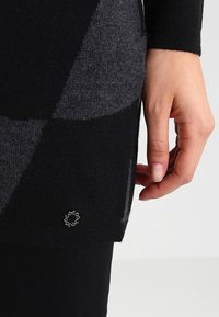 Suéter de punto con un patrón en negro y gris, mostrando una mano en el dobladillo, con un pequeño detalle del logo cerca de la parte inferior, textura suave.