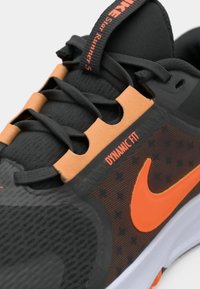 Běžecká obuv Nike s černým síťovaným svrškem, oranžovými akcenty, polstrovaným límcem a texturovanou podrážkou. Obsahuje štítek "DYNAMIC FIT" a systém šněrování.