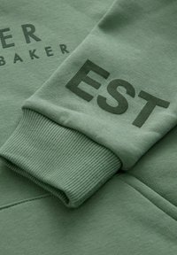 Zelena dukserica s rebrastim rukavima i otisnutim tekstom "BAKER EST." u kontrastnoj nijansi. Mekani materijal s ležernim krojem.