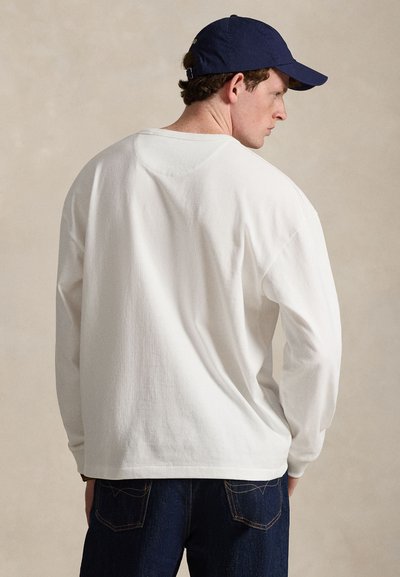 Άνδρας που φοράει λευκό μακρυμάνικο πουκάμισο, σκουρόχρωμο μπλε τζιν και μπλε σκουφί μπέιζμπολ, στραμμένος με την πλάτη σε ένα απλό φόντο.