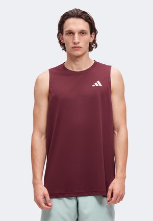 Мъж позира с бордо потник Adidas и светлозелени шорти на изчистен светъл фон.