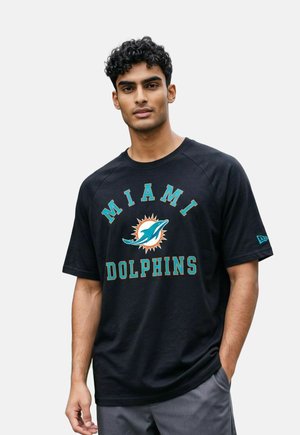Mann mit dunklem lockigem Haar, der ein schwarzes Miami Dolphins T-Shirt und graue Hosen trägt, steht mit einer Hand in der Tasche vor einfarbigem Hintergrund.
