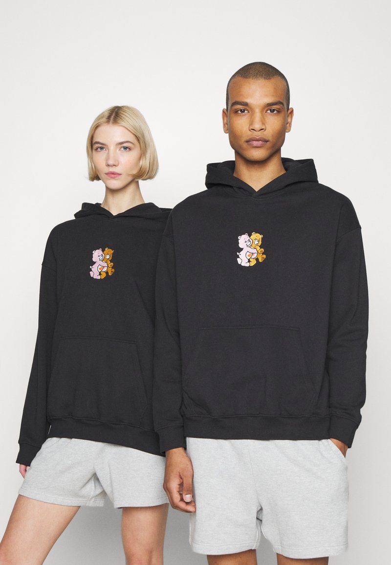 Black hoodies made of cotton blend, featuring a small colorful bear graphic on the front. Models wear gray shorts. Minimalist design.