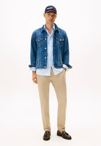 Jonge man met een marineblauwe pet, een blauwe spijkerjas over een lichtblauw gestreept overhemd, beige broek en donkerbruine loafers, staand met handen in de zakken.