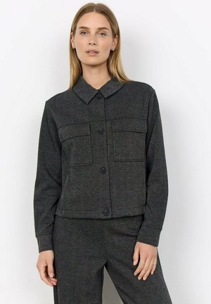 Vrouw met stijl blond haar die een donkergrijs jasje met knopen draagt met twee borstzakken en bijpassende broek tegen een effen witte achtergrond.