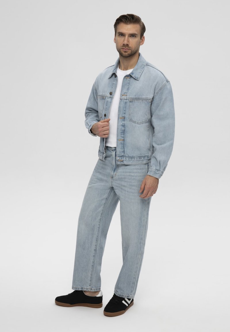 Homme portant une veste en jean bleu clair et un jean avec une chemise blanche et des baskets noires sur un fond clair uni.