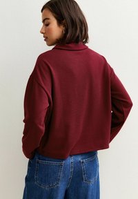 Felpa borgogna corta con colletto alto, maniche larghe e texture morbida, abbinata a jeans in denim blu con tasche posteriori.