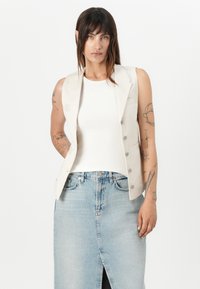 Beige ermeløs vest med en tettsittende design, med en rad med knapper og en lomme. Sammen med en hvit ribbestrikket tanktop og lys blå denimskjørt.