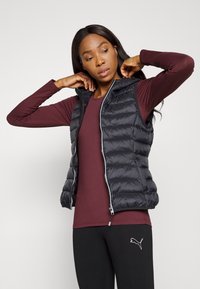 Svart puffer vest med horisontalt quiltet mønster og frontglidelås, båret over en vinrød langermet topp, kombinert med svarte leggings.